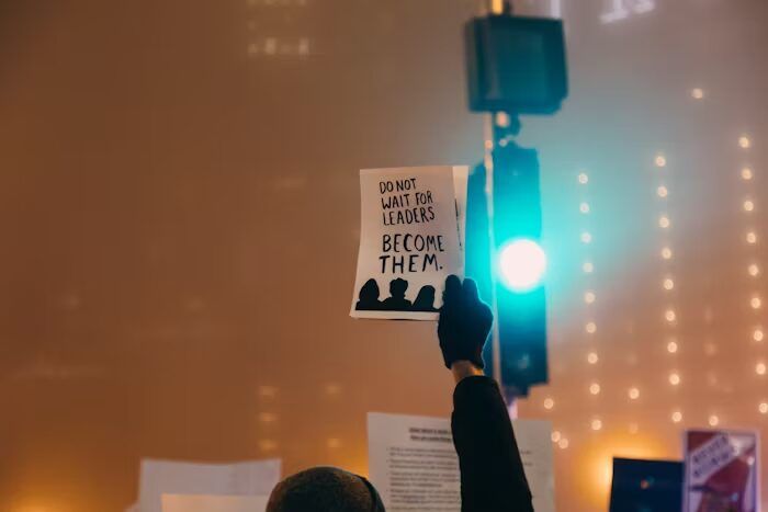 Tangan memegang poster bertuliskan 'DO NOT WAIT FOR LEADERS BECOME THEM.' di malam hari.