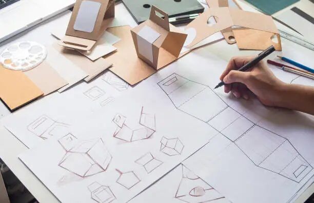 Tangan sedang menggambar sketsa kemasan di atas lembaran kertas, dengan model kemasan berbentuk kotak dan tabung.