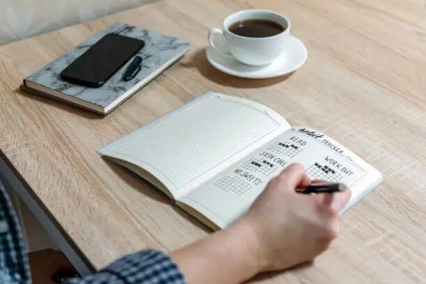 Tangan menulis di buku dengan kopi dan ponsel di meja.