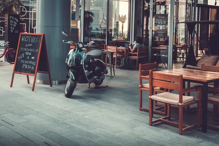 Pemandangan pasar dengan meja dan kursi kayu, motor parkir, dan papan tulis.