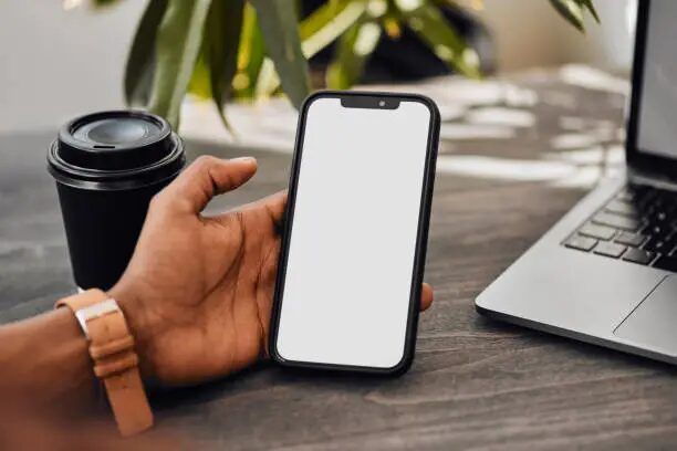 Tangan memegang smartphone putih di meja dengan laptop dan kopi di latar belakang.