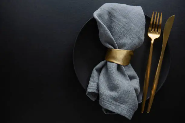 Tata letak peralatan makan elegan dengan piring hitam, garpu emas, dan kain napkin abu-abu.
