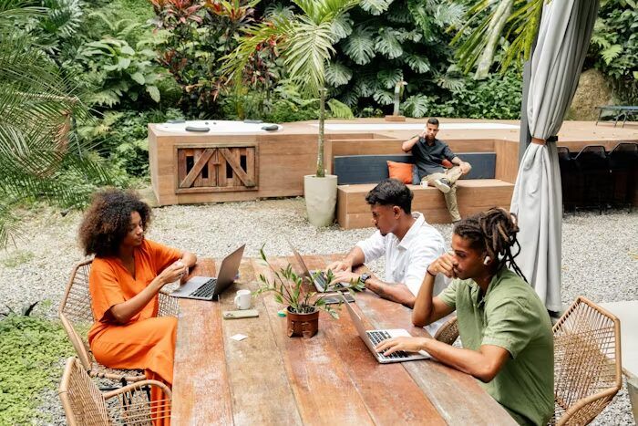 Tiga orang sedang bekerja dengan laptop di meja di luar ruangan dengan tanaman dan bangunan modern di latar belakang.