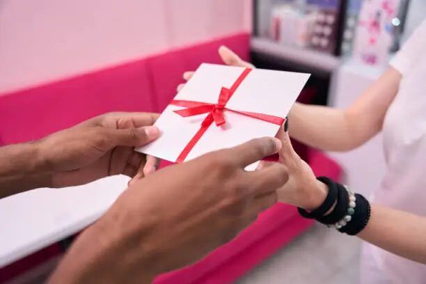 Tangan memegang kotak kecil berwarna putih dengan tali merah, di depan meja berwarna pink.