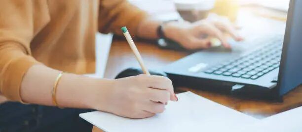Tangan sedang menulis di kertas dengan laptop di depannya.