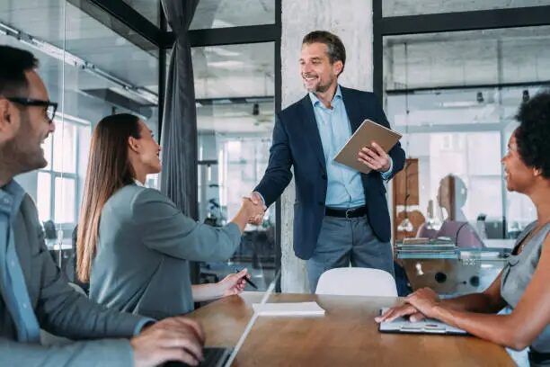 Dua orang sedang berjabat tangan di meja rapat, latar belakang kantor modern.