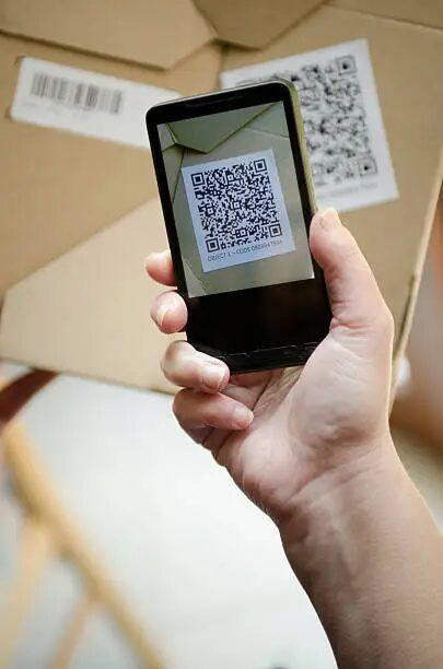 Tangan memegang smartphone dengan QR code di layar, latar belakang kardus.