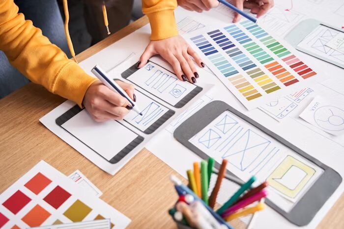 Tangan memegang pensil dan sketsa layar mobile dengan warna-warna cerah di meja.