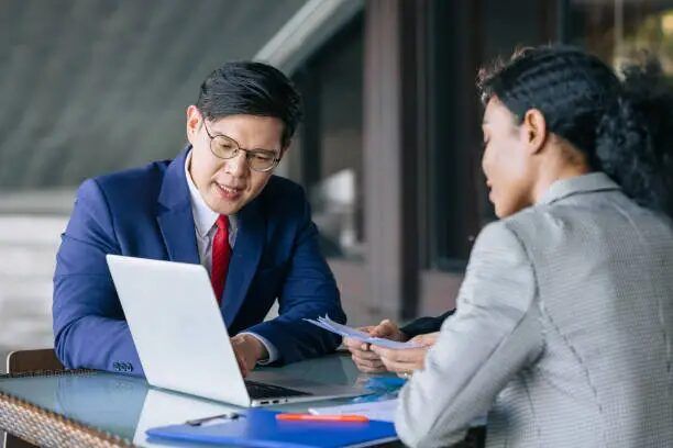 Dua orang berbincang di meja dengan laptop, setting bisnis.