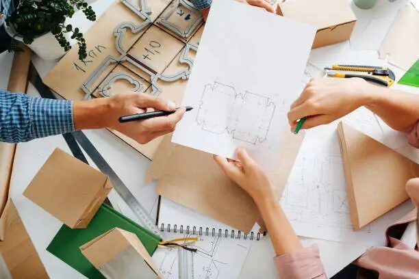 Tiga orang sedang merancang desain grafis di meja kerja dengan alat tulis dan sketsa.