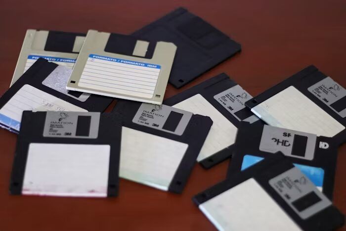 Koleksi floppy disk berbagai ukuran dan kondisi di meja kayu.