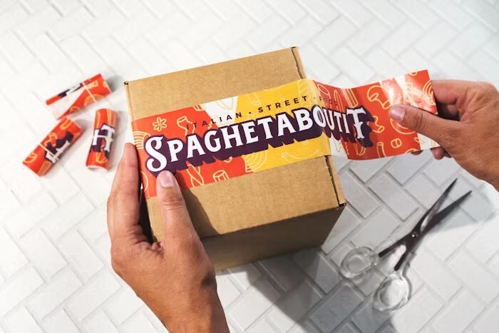 Tangan memegang kotak karton berlabel Spaghetaboutif di samping alat pemotong dan stiker.