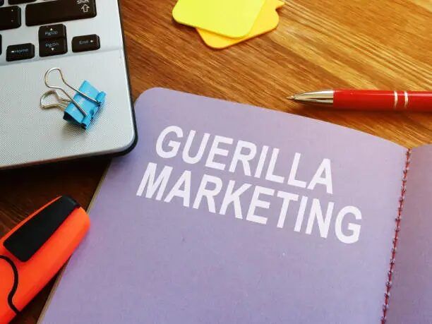 Notebook bertuliskan 'GUERILLA MARKETING' dengan alat tulis dan laptop di sekitarnya.
