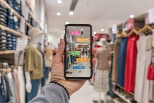 Pelanggan memegang smartphone di depan rak pakaian sebagai ilustrasi promosi retail yang terhubung antara toko fisik dan digital