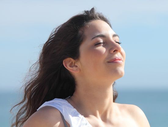Wanita dengan rambut panjang menikmati angin di pantai dengan latar laut biru.