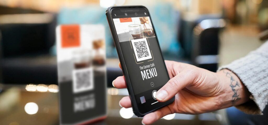 Tangan memegang smartphone dengan aplikasi menu cafe dan QR code.