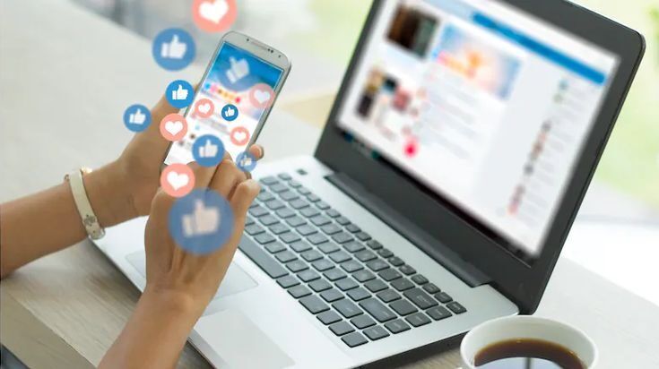 Tangan memegang smartphone dengan ikon media sosial di layar, laptop di belakangnya, dan kopi di samping.