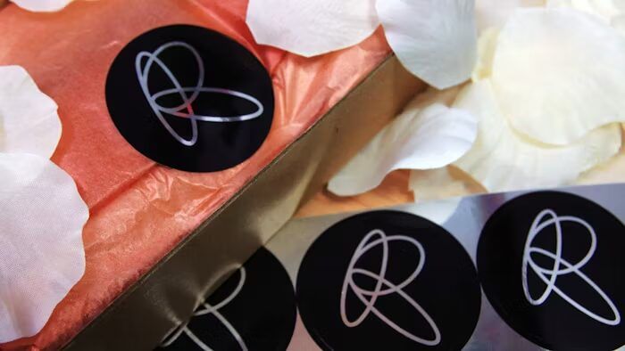 Stiker hitam dengan logo putih di atas kain oranye dan daun bunga putih.