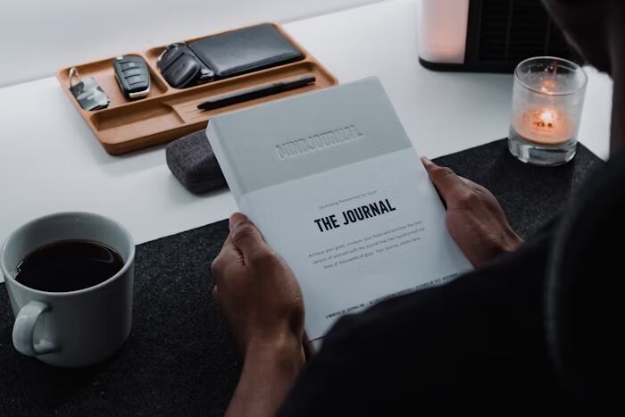 Tangan memegang buku journal di meja kerja dengan kopi dan aksesori.