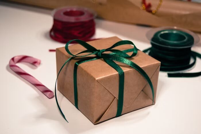 Paket hadiah berwarna coklat dengan pita hijau, disertai permen cokelat dan benang pita.