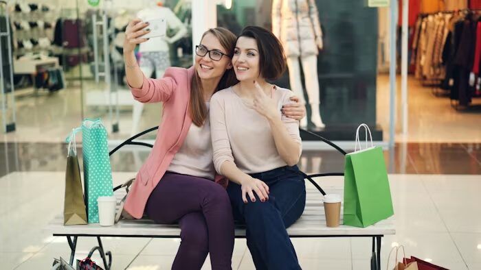 Dua wanita sedang selfie di tempat nongkrong di mall dengan belanjaan di sekitar mereka.