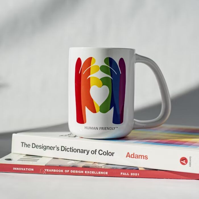Mug putih dengan desain warna-warni dan tulisan 'HUMAN FRIENDLY™' di atas buku 'The Designer’s Dictionary of Color'.