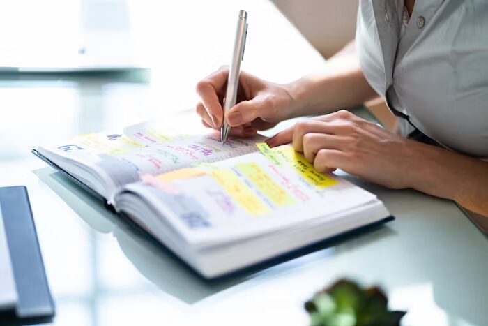 Tangan menulis di buku agenda dengan sticky notes warna-warni.