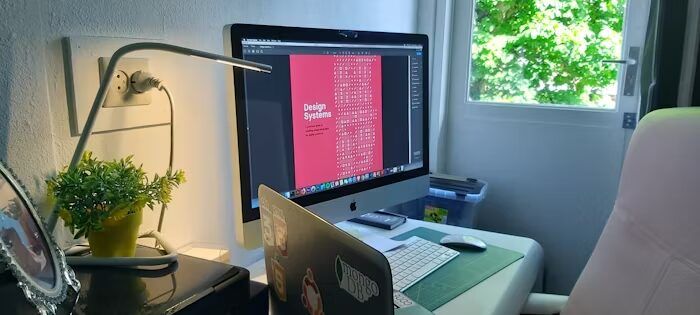 Meja kerja dengan komputer dan laptop yang menunjukkan proses memilih tipografi untuk desain bahan promosi cetak.