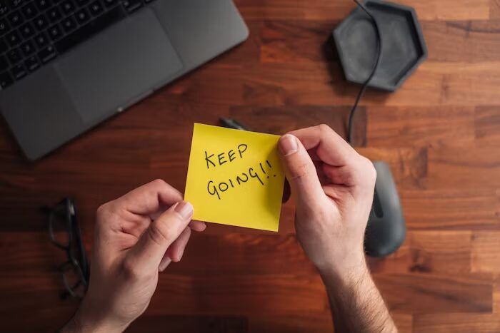 Tangan memegang stiker kuning dengan tulisan 'Keep Going!!' di meja kerja.