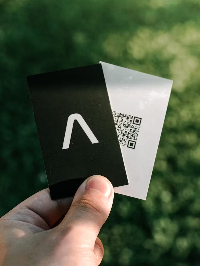 Tangan memegang dua kartu dengan logo dan QR code, latar hijau alami.