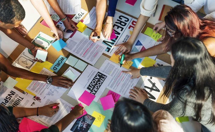 Grup orang sedang bekerja sama dalam sesi desain grafis di meja penuh dengan poster dan stiker warna-warni.