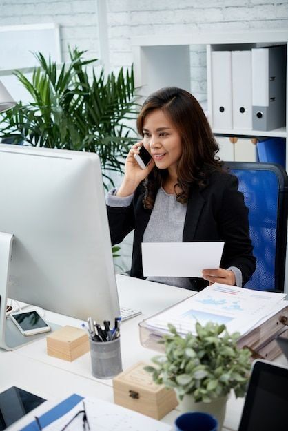 Seorang wanita sedang bekerja di meja kantor dengan komputer dan handphone di tangan.