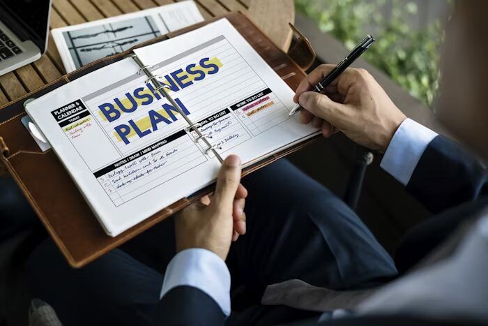 Tangan menulis pada lembaran bisnis plan di Uprint.id saat menyiapkan konsep booklet landscape A5 untuk kebutuhan bisnis
