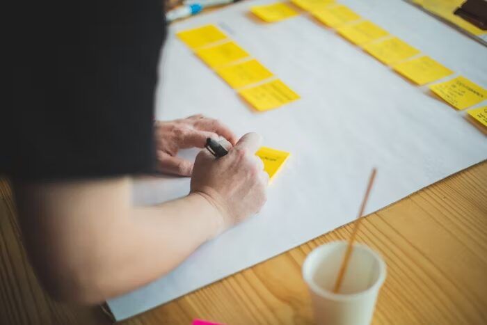 Tangan memegang marker dan menulis di atas lembaran besar kertas dengan sticky notes kuning untuk menyusun layout booklet landscape