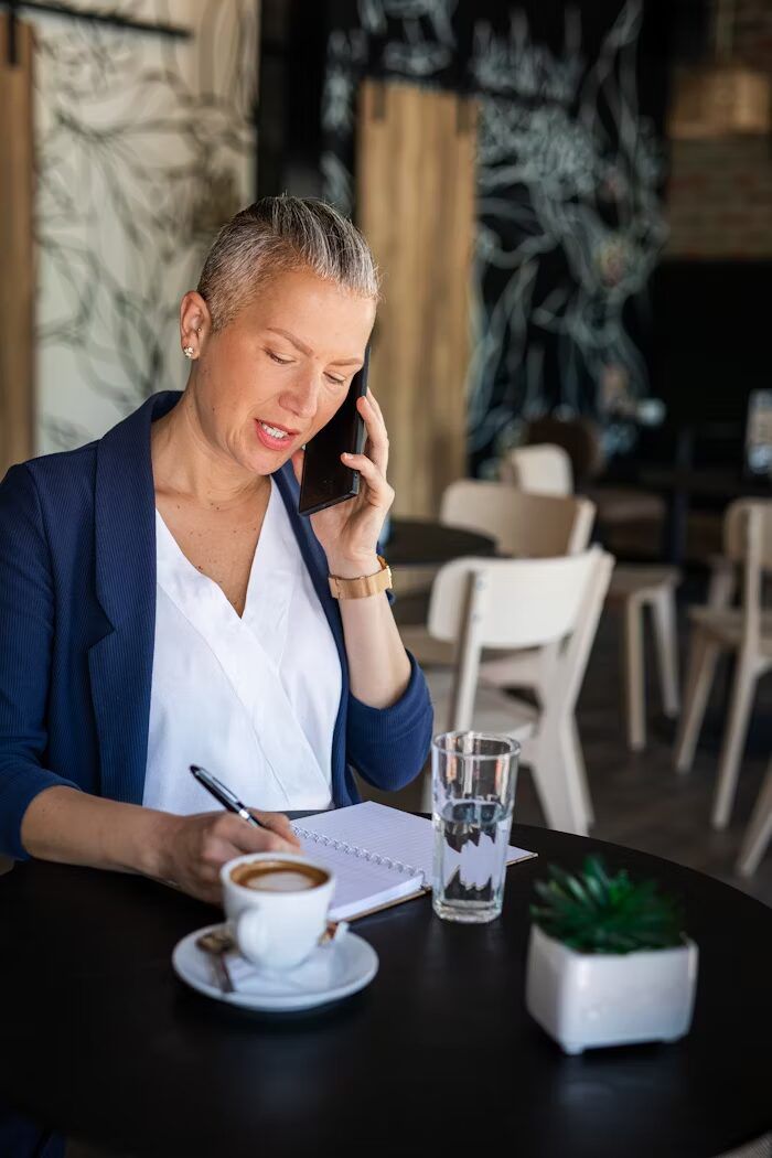 Perempuan berbaju kerja sedang berbicara telepon sambil menulis di cafe.