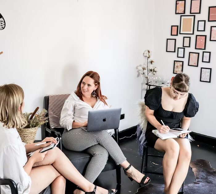 Tiga wanita sedang berbicara dan bekerja di ruang kerja minimalis dengan laptop dan catatan.