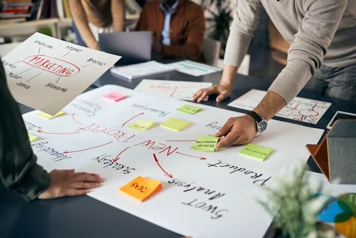 Tiga orang sedang merencanakan strategi marketing dengan papan tulis dan post-it warna-warni.