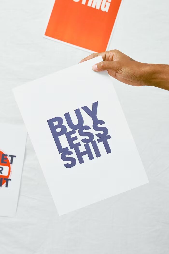 Tangan memegang poster putih dengan pesan 'Buy Less Shit' dalam huruf besar.