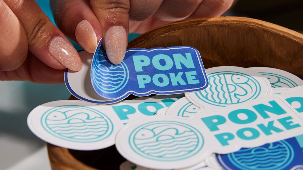 Tangan memegang stiker Pon Poke biru dan putih di atas wadah kayu.