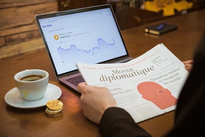 Tangan memegang majalah 'Monde diplomatique' di meja cafe dengan laptop dan kopi.