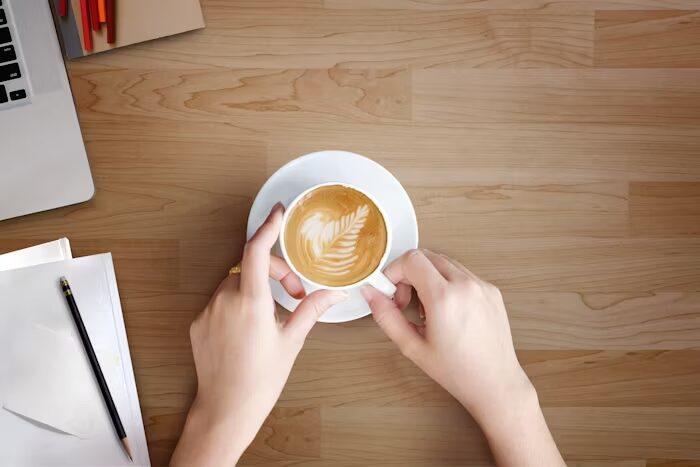 Tangan memegang cangkir kopi dengan seni latte art di atas meja kayu.