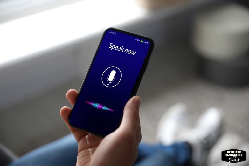 Tangan memegang smartphone dengan layar menampilkan fitur voice recognition.