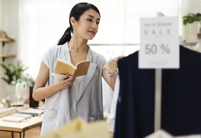 Perempuan membaca buku sambil memilih pakaian di toko dengan penawaran diskon 50%.
