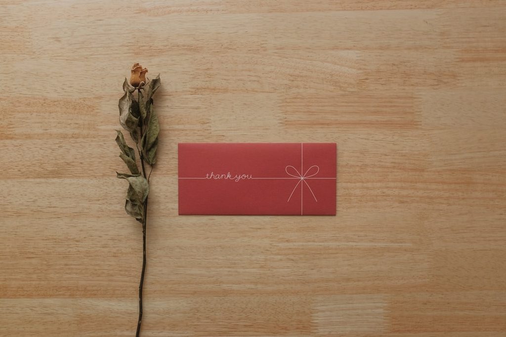 Kartu terima kasih merah dengan tulisan 'thank you' dan ikat bunga di atas meja kayu.