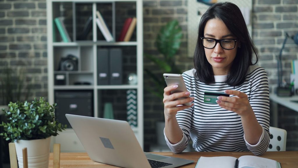 Perempuan memegang kartu kredit dan smartphone, sedang berada di depan laptop di ruang kerja.