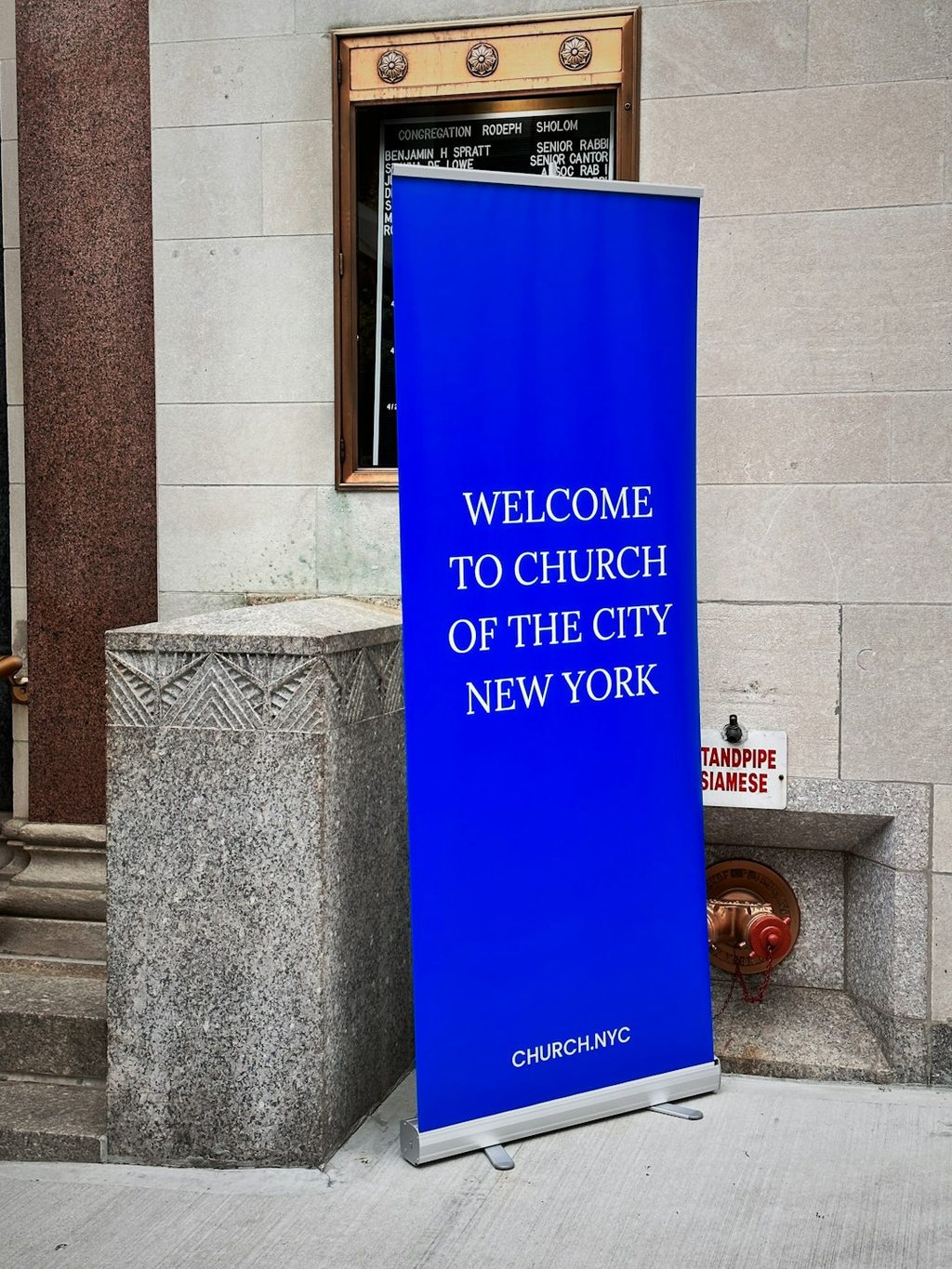 Banner biru dengan tulisan 'Welcome to Church of the City New York' di depan gedung.