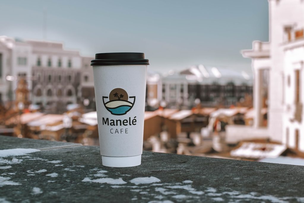 Cangkir kopi putih dengan logo Manelé Cafe di latar bangunan kota.