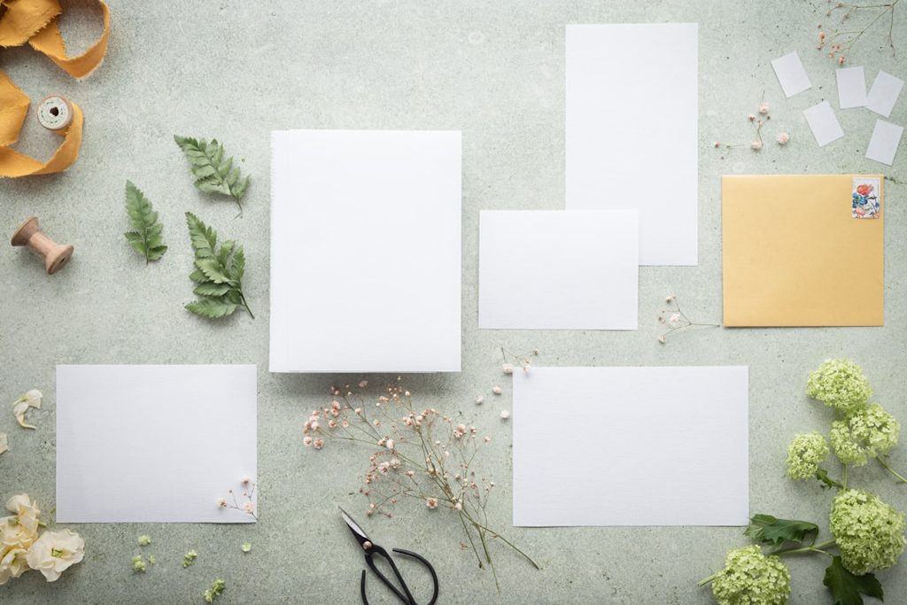 Gambar menunjukkan beberapa lembar kertas putih dan bunga di meja, ideal untuk contoh desain percetakan.