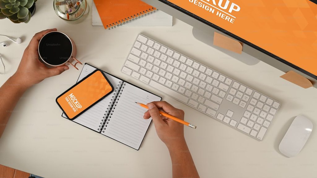 Tangan menulis di buku notepad dengan smartphone dan komputer di dekatnya.