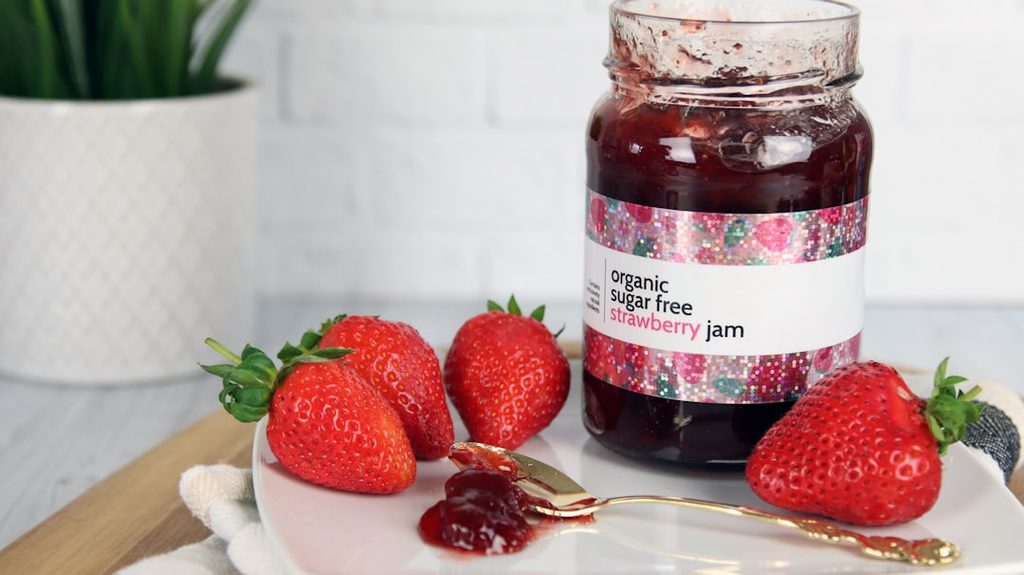 Botol jam strawberry organik tanpa gula dengan stiker label dan buah strawberry segar di sekitarnya.
